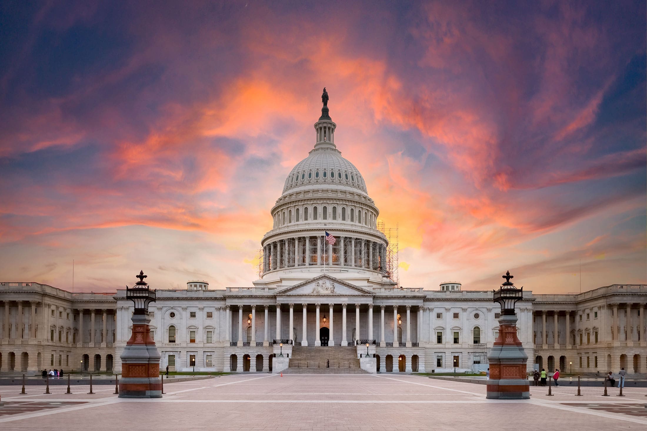 us-capitol-building-dramatic-sunset-sita. Front facade of the United States Capitol building at sunset with a dramatic orange and purple sky, an American flag visible beneath the dome
