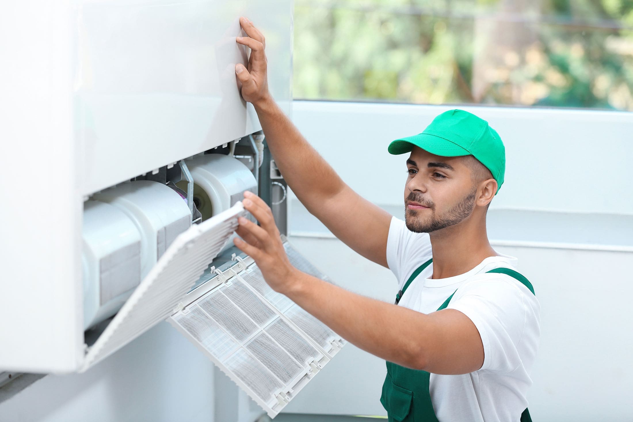 technician-servicing-mini-split-air-conditioner-sita. HVAC technician in a green uniform and cap opening the filter panel of a wall-mounted mini split air conditioning unit