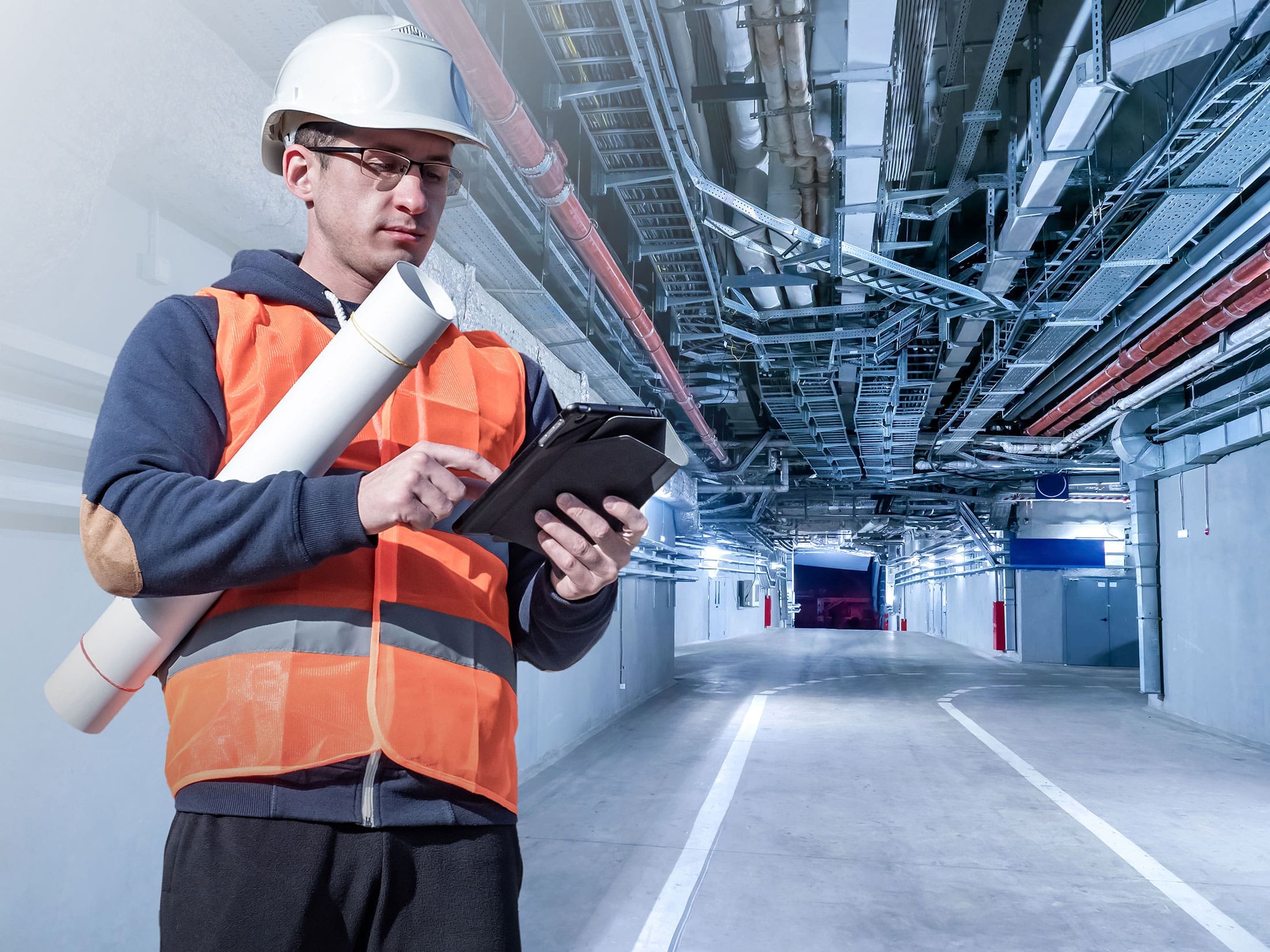 engineer-tablet-underground-mechanical-corridor-sita. Engineer in an orange safety vest and white hard hat reviewing a tablet while holding rolled blueprints in a underground mechanical corridor