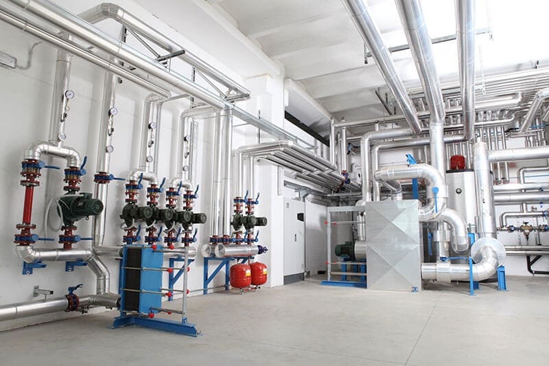 mechanical-room-red-valve-manifolds-blue-pipe-supports-sita. Bright commercial mechanical room with silver insulated piping, red motorized valve manifolds, blue expansion tanks, and a large air handling unit under a skylit white ceiling