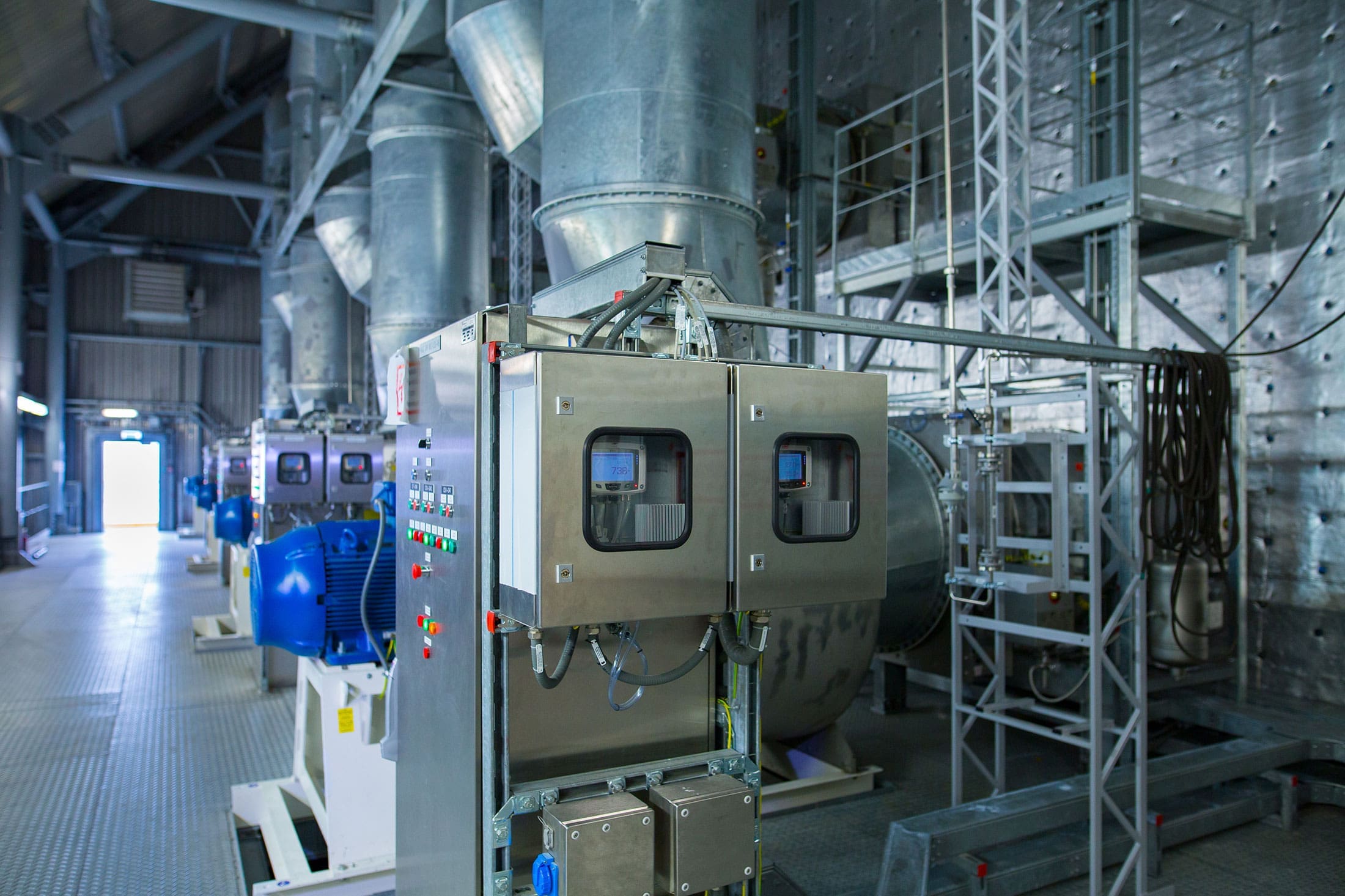 industrial-control-panel-ventilation-equipment-sita. Stainless steel industrial control panel with digital displays mounted beneath large galvanized ventilation ducts inside a large manufacturing facility