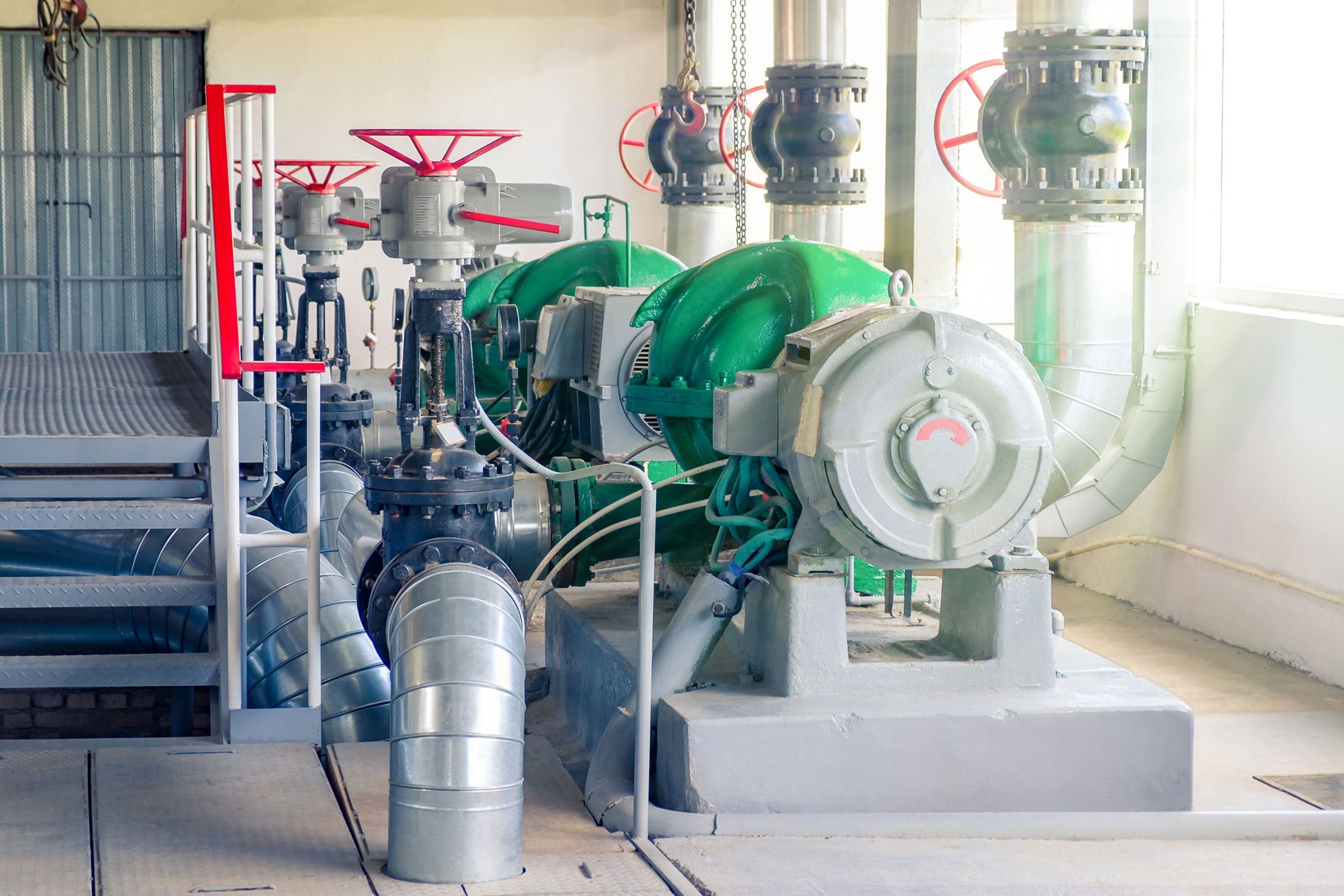 industrial-pump-room-green-motors-red-valves-sita. Industrial pump room with large green centrifugal pumps, red gate valves, flanged piping, and a silver duct on a concrete floor