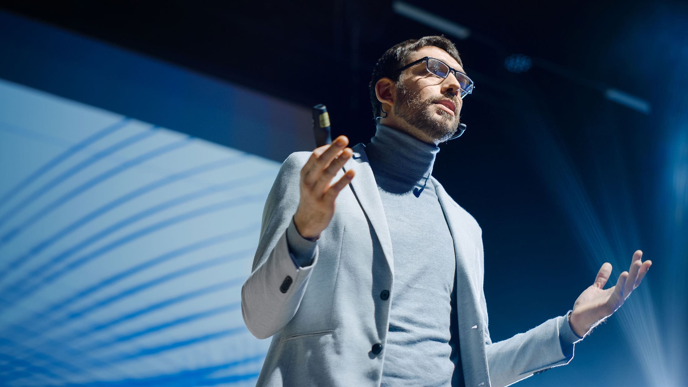 professional-speaker-stage-blue-background-sita. A bearded male speaker in a gray blazer and turtleneck gestures expressively on a darkened stage in front of a blue abstract projection screen