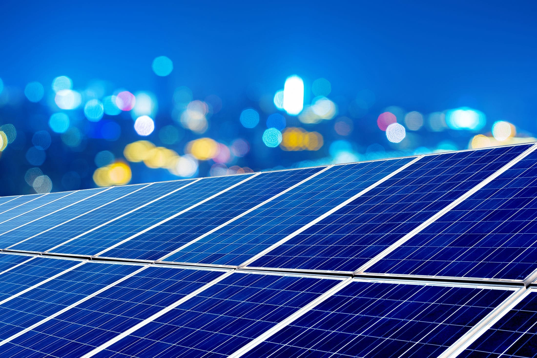solar-panels-night-city-bokeh-sita. Close-up of blue solar panels at night with blurred colorful city lights in the background creating a bokeh effect