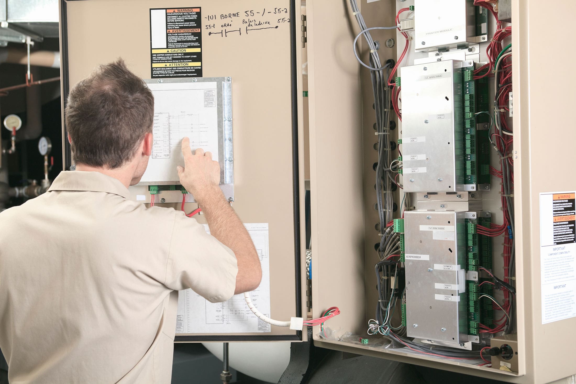 technician-pointing-hvac-control-panel-schematic-sita. Rear view of a technician in a beige shirt pointing at a wiring schematic on an open HVAC control panel packed with labeled control modules and colored wiring