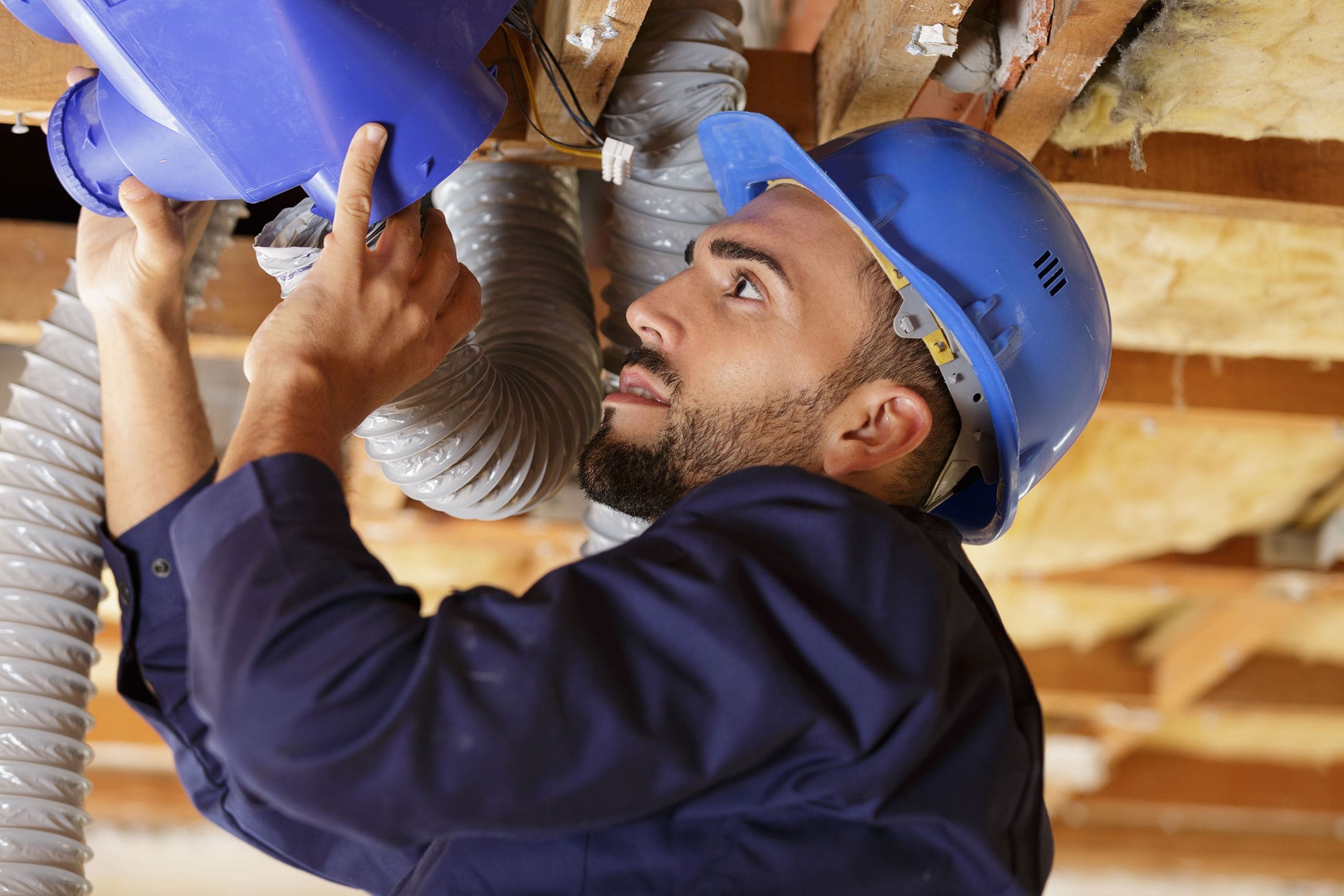 technician-installing-flexible-ductwork-attic-sita. A technician in a blue hard hat looking upward while connecting blue and flexible gray ductwork inside a wood-framed attic space with insulation visible