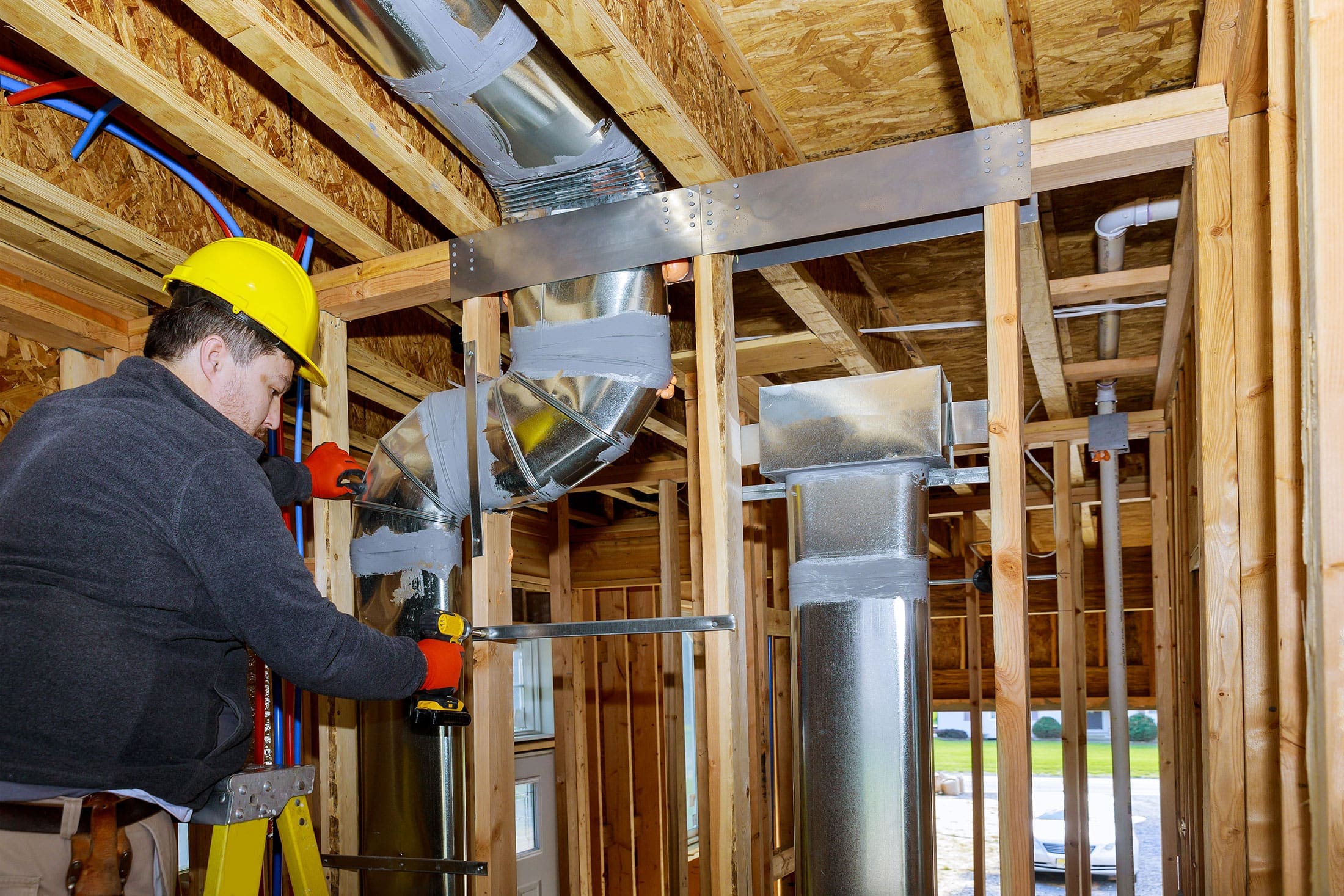 hvac-duct-installation-new-construction-framing-sita. Worker in a yellow hard hat and red gloves installing silver HVAC ductwork inside a wood-framed building under construction