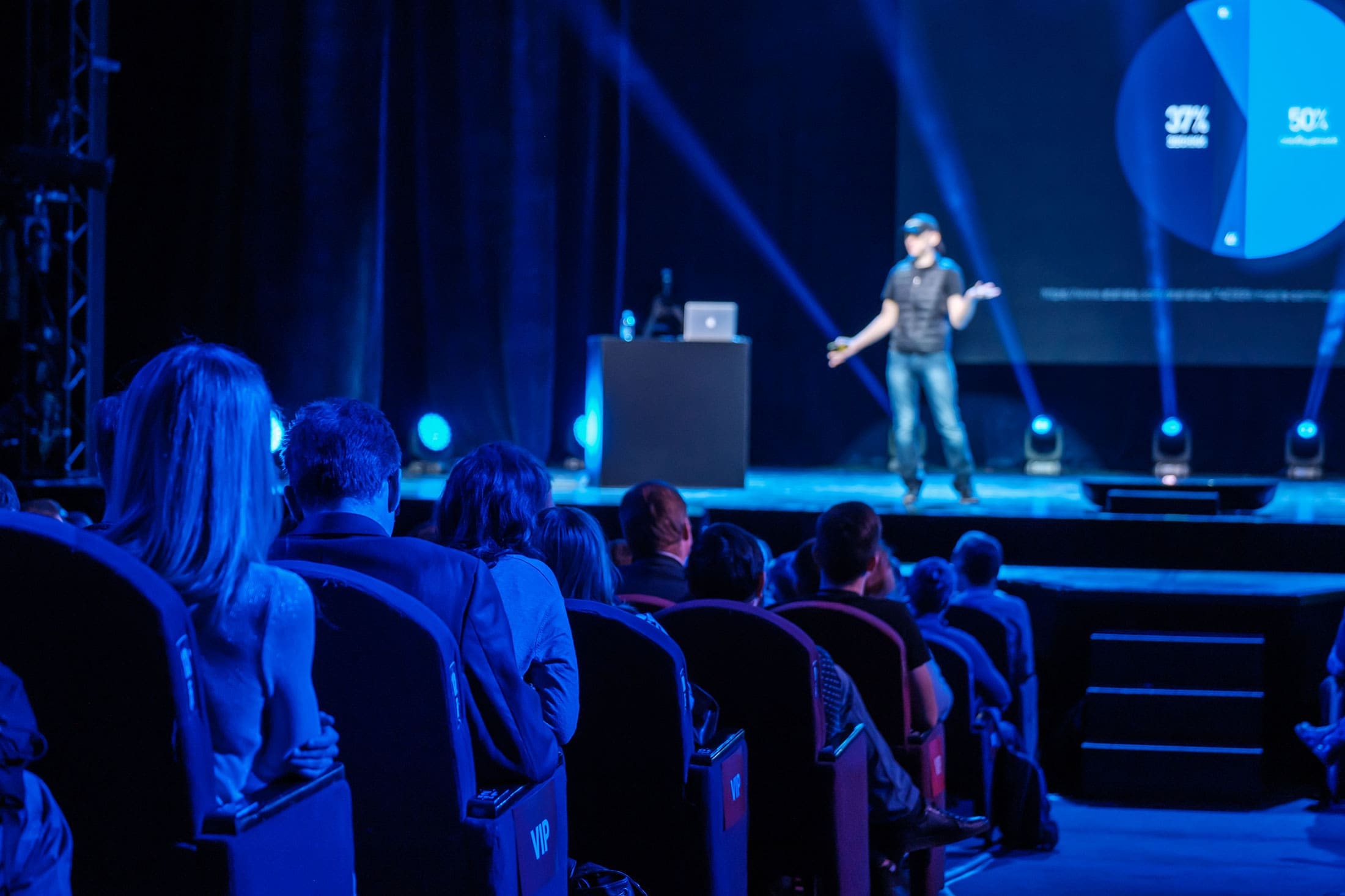 vip-conference-audience-stage-presenter-blue-lighting-sita. Rear view of a VIP audience in theater-style seating watching a presenter on a dramatically lit blue stage with a pie chart slide on the large screen