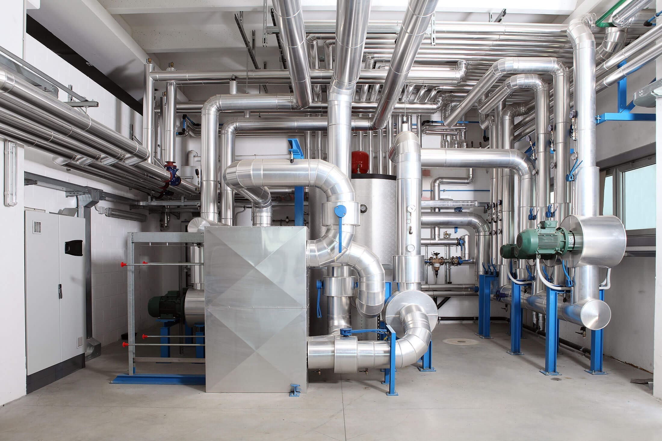 commercial-mechanical-room-silver-piping-blue-supports-sita. Well-organized commercial mechanical room with an extensive network of silver insulated pipes, blue steel supports, green pump motors, and a large rectangular air handler unit