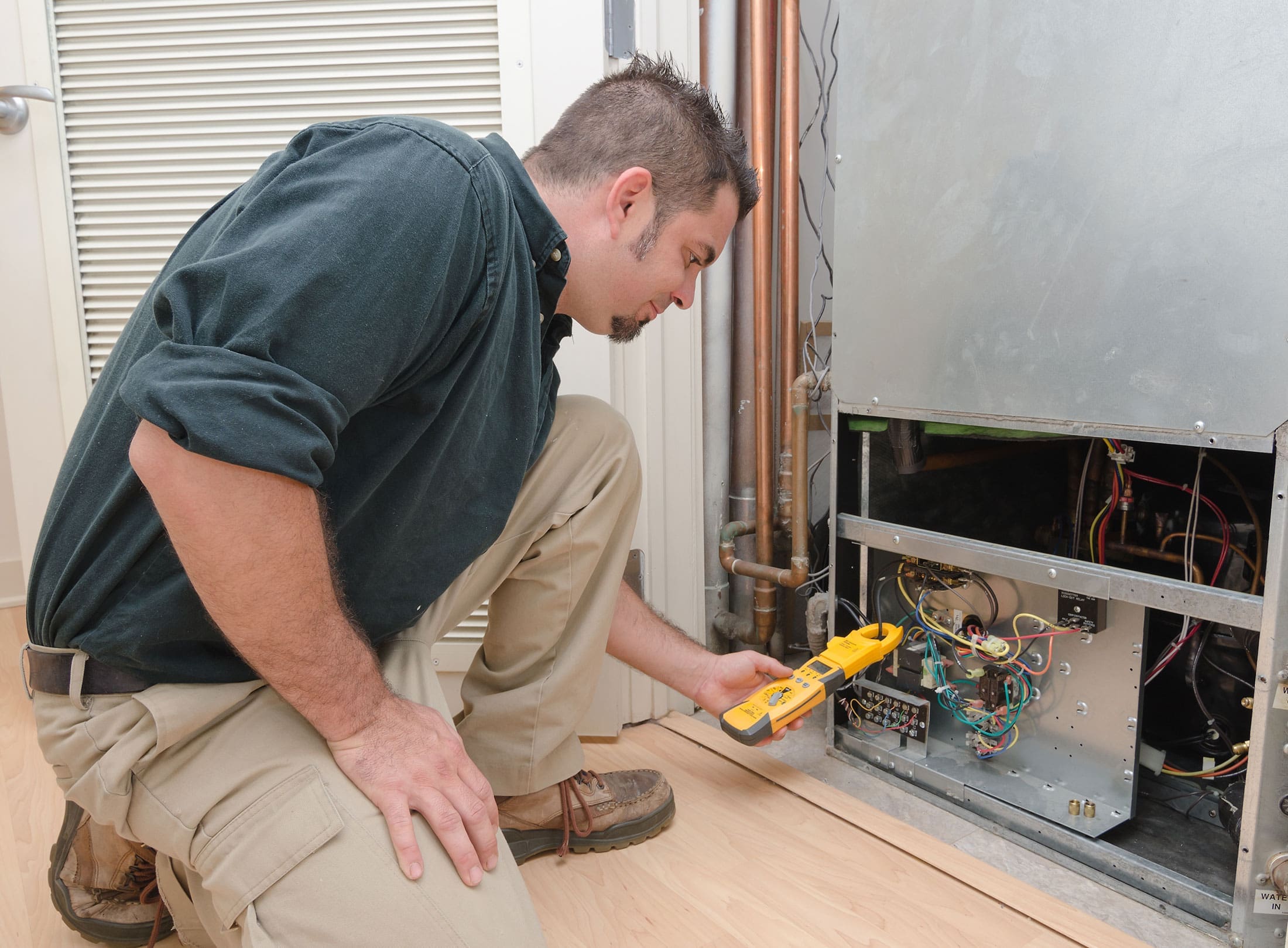 hvac-technician-testing-residential-air-handler-sita. HVAC technician crouching beside an open residential air handler unit using a yellow clamp meter to test internal wiring and components