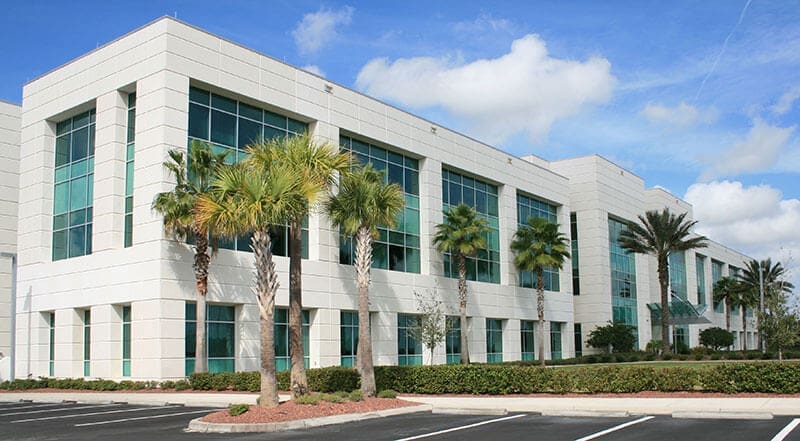 two-story-florida-commercial-office-building-palm-trees-sita. Exterior view of a two-story beige commercial office building with teal-tinted windows, palm trees in the parking lot landscape, and a partly cloudy blue sky