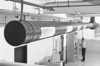 large-round-ventilation-duct-mechanical-room-sita. A large horizontal round black and silver ventilation duct suspended from the ceiling of a white industrial mechanical room with a person visible in the background