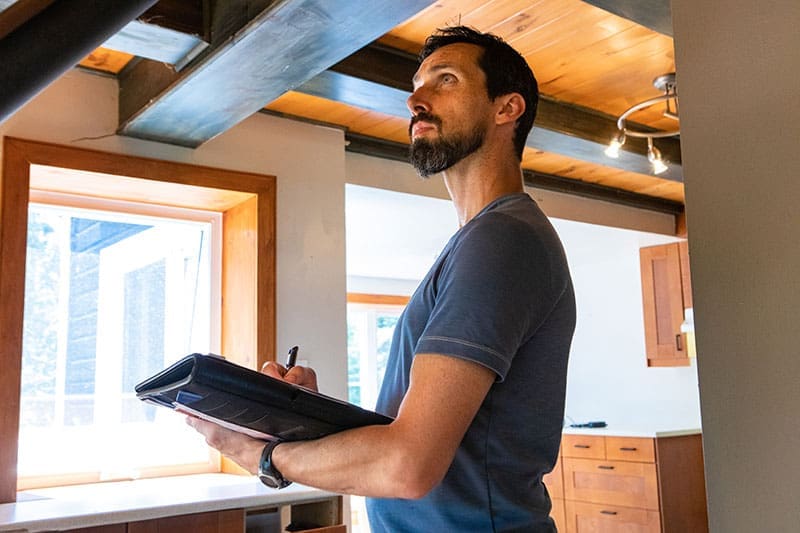 home-inspector-writing-notes-ceiling-inspection-sita. Bearded male inspector in a blue t-shirt holding a notepad and pen while looking up at exposed wood ceiling beams inside a residential home