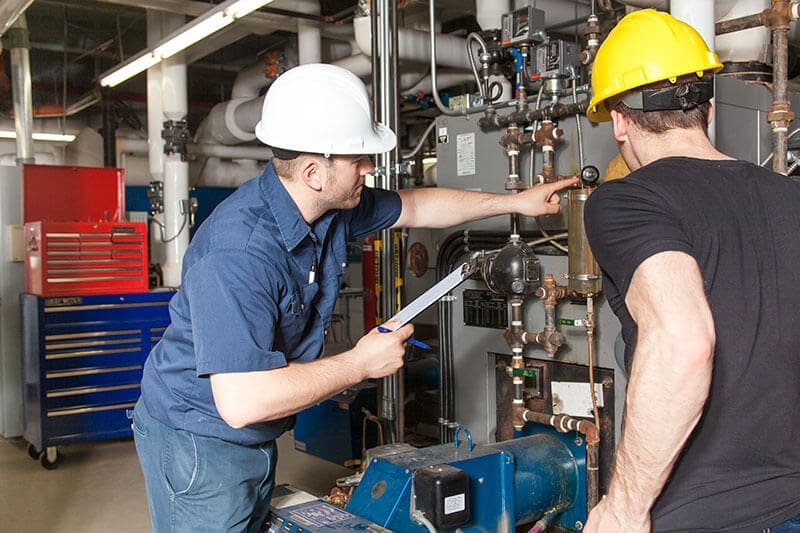 two-technicians-inspecting-mechanical-equipment-clipboard-sita. Two technicians in white and yellow hard hats examining piping and equipment in a mechanical room while one holds a clipboard and points at a component