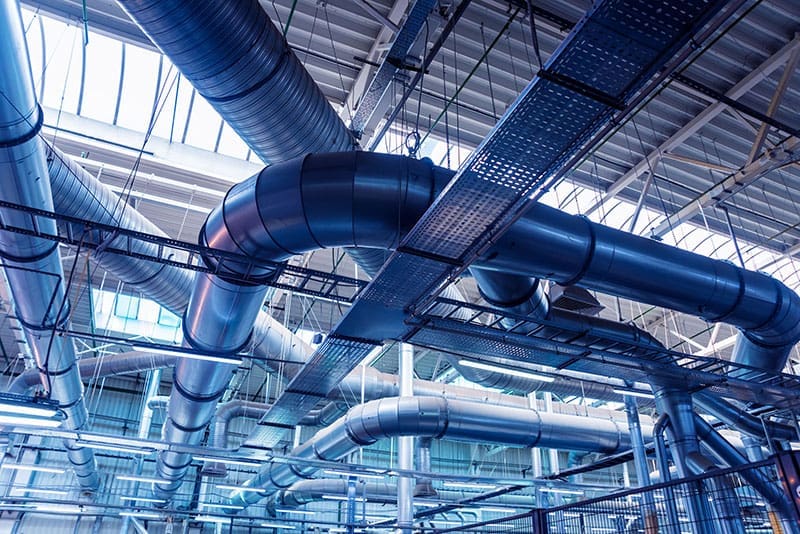 industrial-hvac-ductwork-ceiling-sita. Large silver industrial HVAC ductwork and pipe systems suspended from a high ceiling in a commercial facility bathed in cool blue light