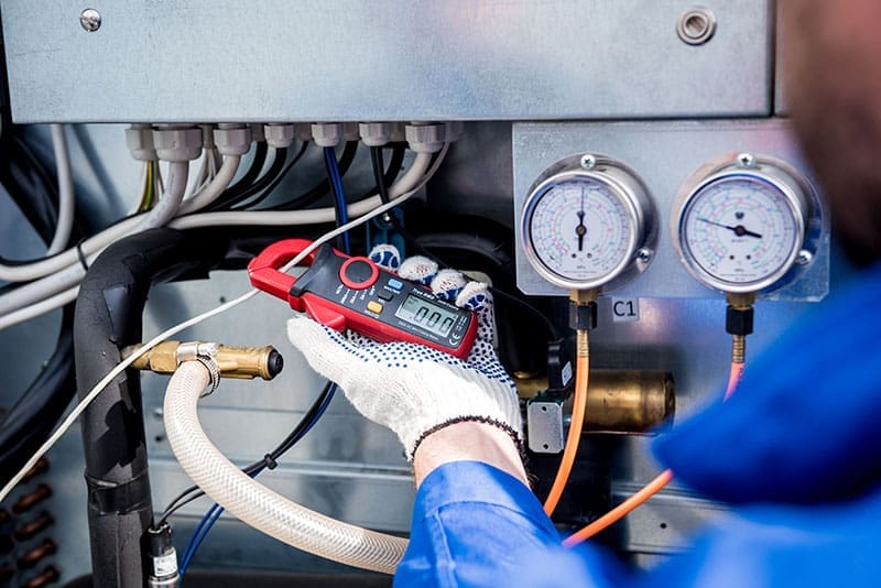 hvac-technician-clamp-meter-diagnostic-sita. HVAC technician in blue uniform and work gloves using a red clamp meter on equipment with pressure gauges visible in the background