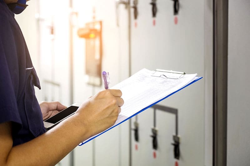 electrical-panel-inspection-clipboard-sita. A technician in a dark uniform writing on a clipboard while standing in front of a row of gray electrical control panels