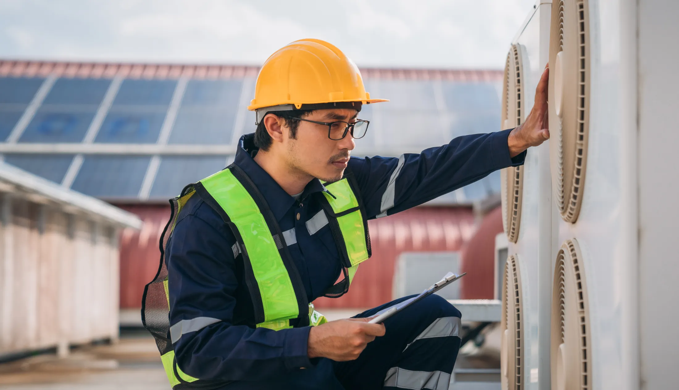hvac-technician-inspecting-rooftop-cooling-system-sita. HVAC technician in safety gear inspecting an outdoor cooling unit while holding a clipboard on a rooftop installation site.