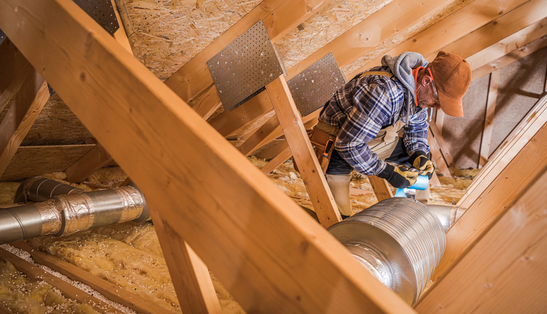 hvac-technician-installing-attic-ductwork-sita. HVAC technician working in an attic space installing and sealing residential air ductwork between wooden roof beams.