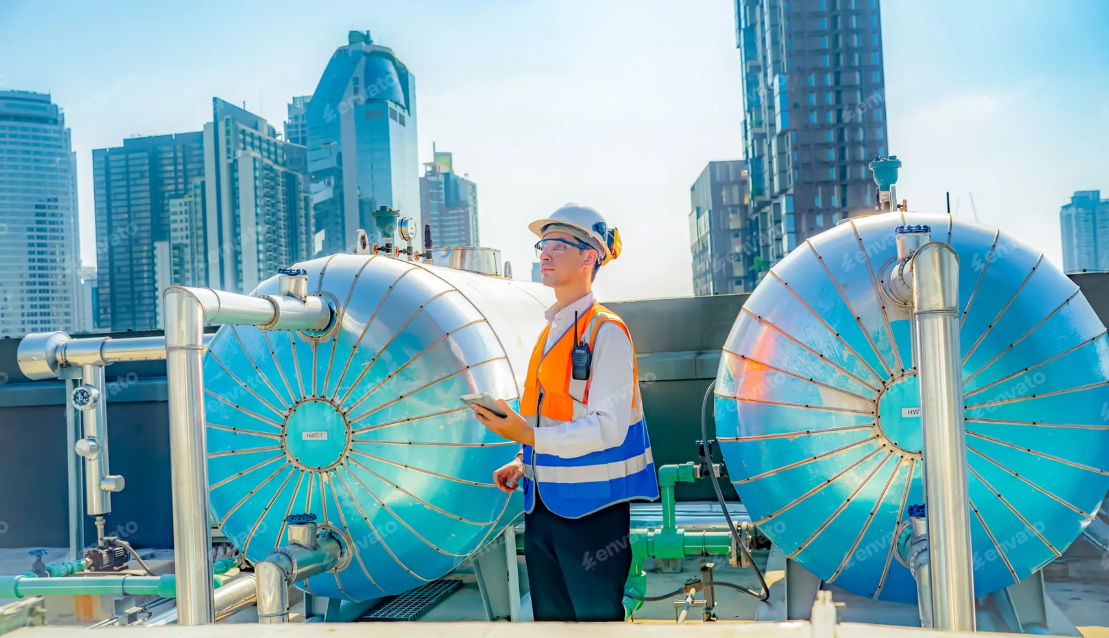 hvac-engineer-inspecting-industrial-rooftop-systems-sita. HVAC engineer standing on a rooftop mechanical site inspecting large industrial pressure tanks and piping systems in an urban environment.