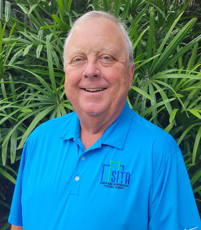 greg-driggers-senior-project-analyst-sita. Portrait of Greg Driggers, Senior Project Analyst at SITA, wearing a blue company polo shirt and smiling outdoors in front of tropical greenery.