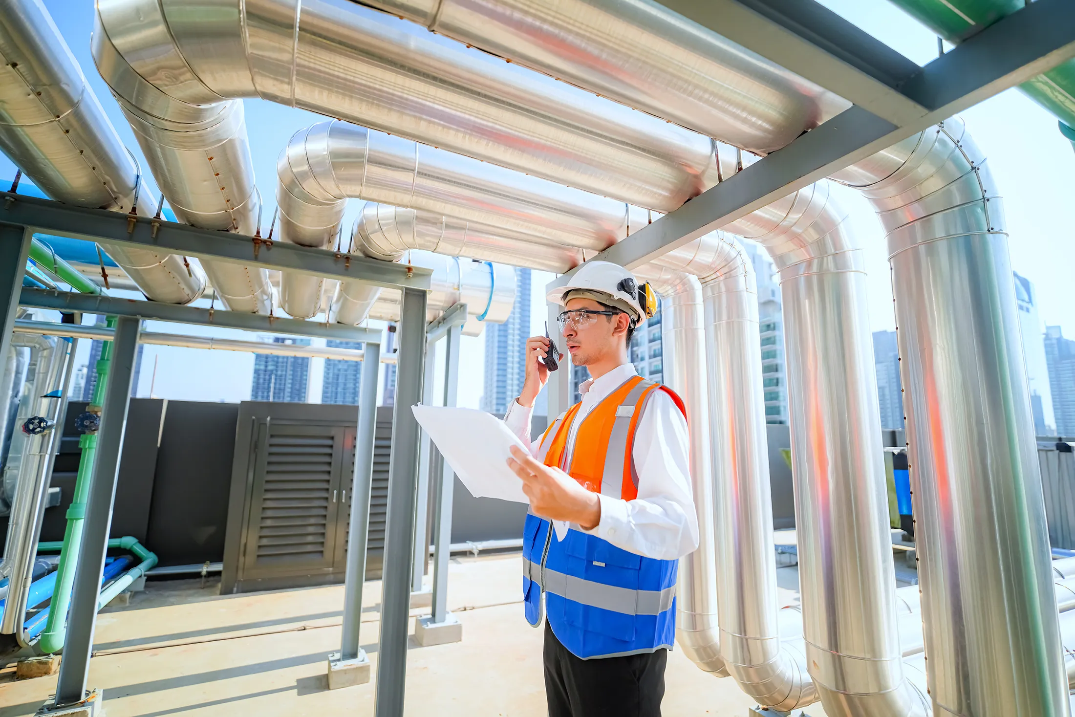 engineer-inspecting-industrial-hvac-and-piping-system-sita. Engineer wearing safety gear inspecting industrial ventilation pipes and HVAC systems while holding project documents at a facility site.