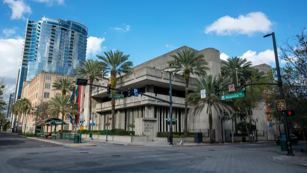 orlando-public-library-and-downtown-cityscape-sita. Urban city street featuring the Orlando Public Library building surrounded by palm trees, traffic lights, and nearby modern high-rise architecture under a blue sky.