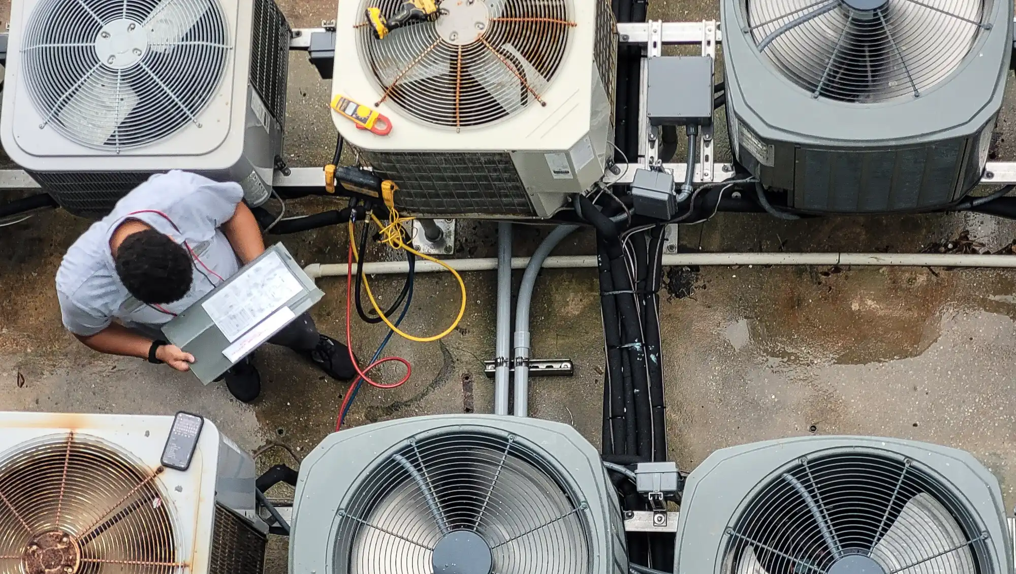 HVAC technician overhead view testing and measuring multiple outdoor air conditioning condenser units with diagnostic equipment and electrical cables.