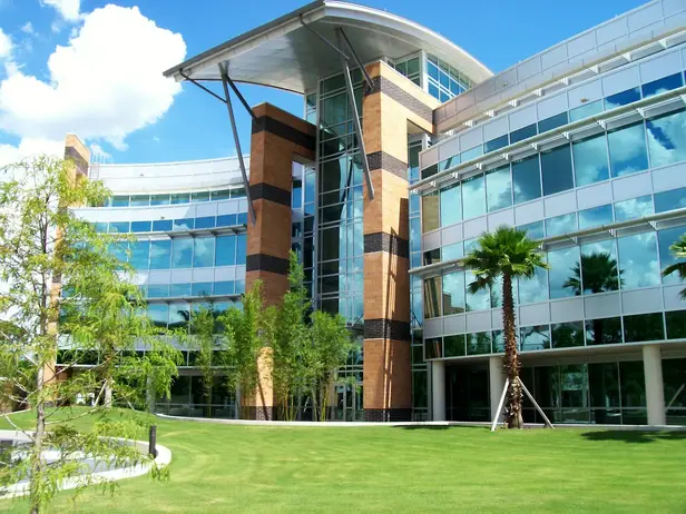 modern-glass-office-building-with-landscaped-campus-sita. Modern multi-story glass office building with curved architectural features, large windows, palm trees, and a landscaped green lawn under a bright blue sky.