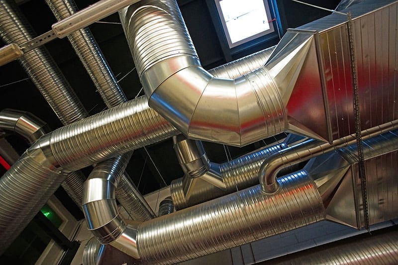 silver-hvac-ductwork-junction-commercial-ceiling-sita. Upward view of polished silver round and rectangular HVAC ducts intersecting at a ceiling junction in a dark commercial space with red accent lighting visible in the background