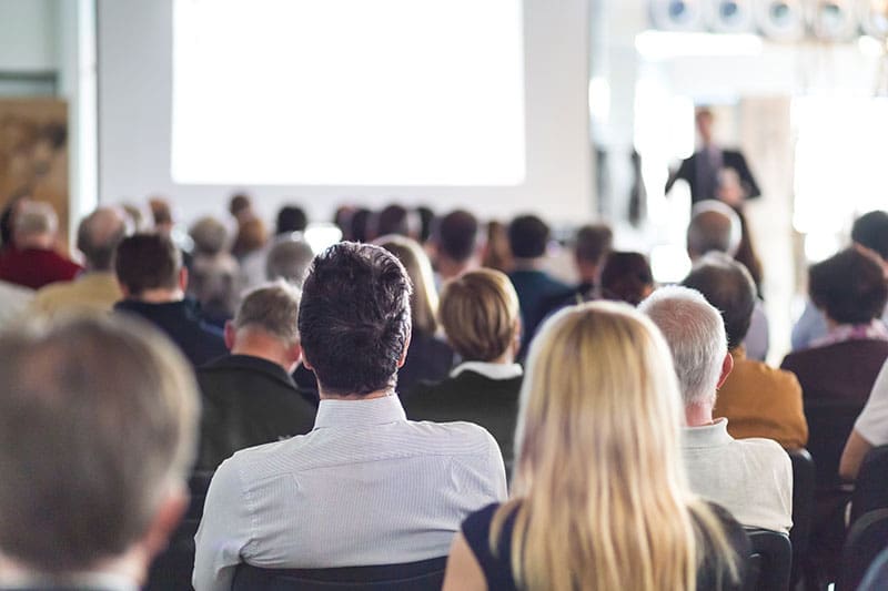 business-conference-audience-presenter-sita. Rear view of a large seated audience facing a presenter at the front of a bright conference room with a projection screen