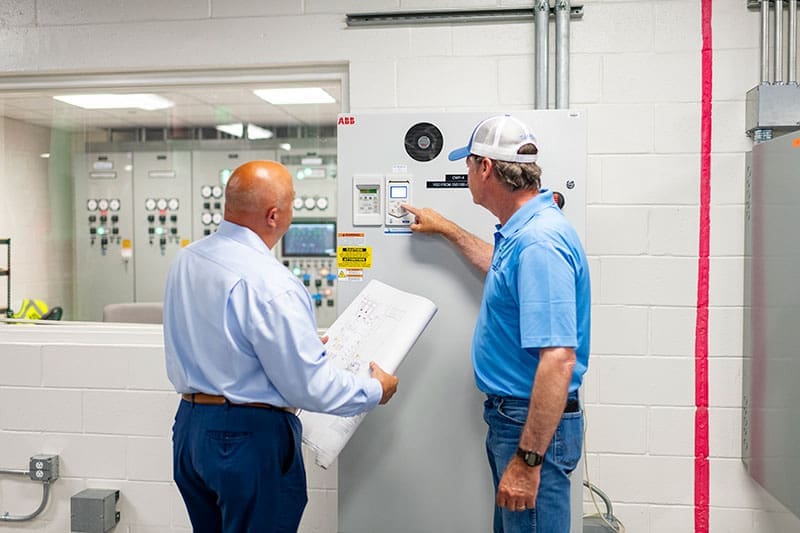 two-men-inspecting-abb-electrical-control-panel-mechanical-room-sita. Two men that work at SITA are working on proper installations of building HVAC Commissioning