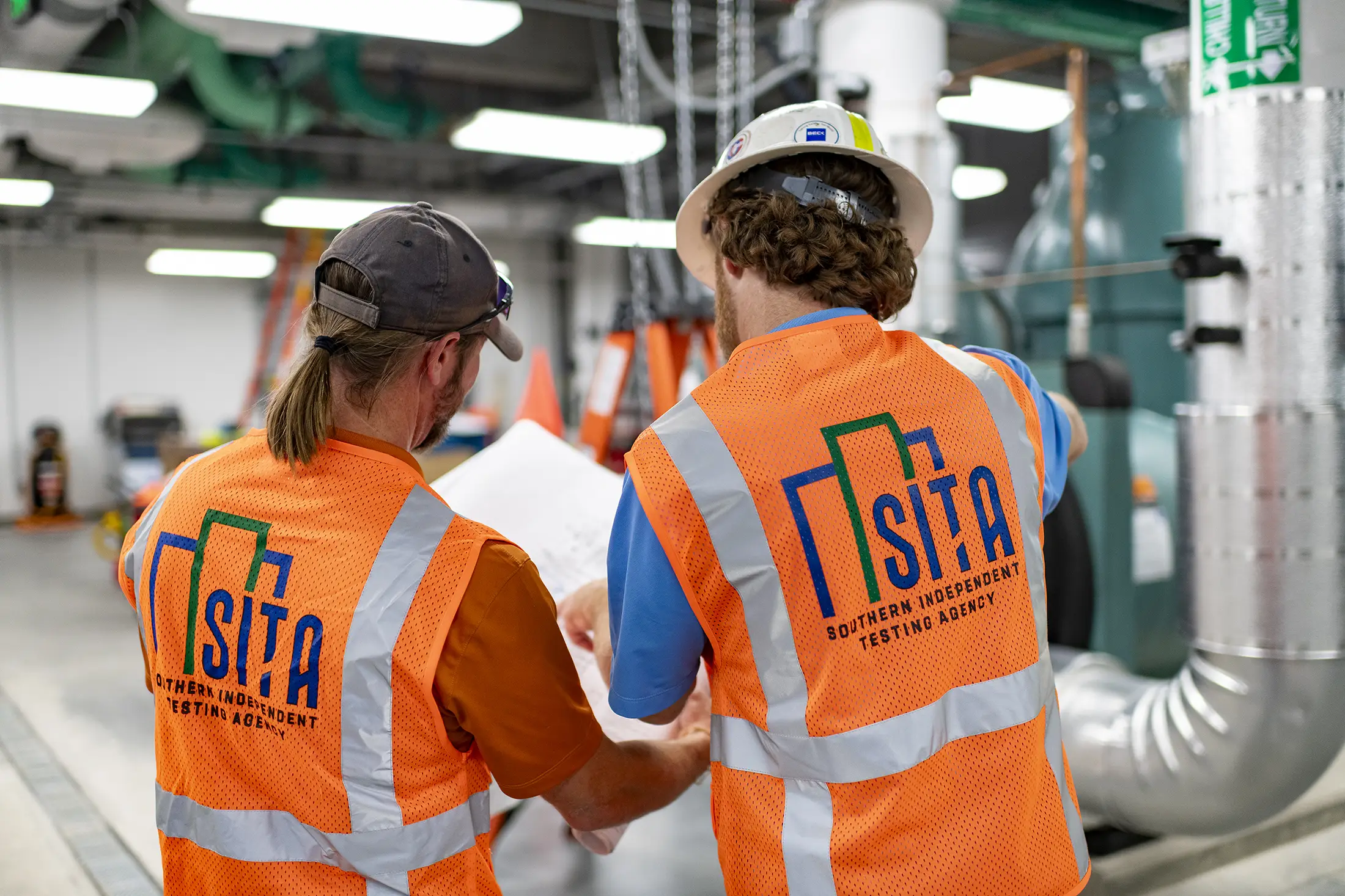 sita-hvac-testing-balancing-plan-review-tampa-florida-sita. Two SITA technical employees reviewing HVAC system plans inside a commercial mechanical room during testing and balancing operations in Tampa, Florida.