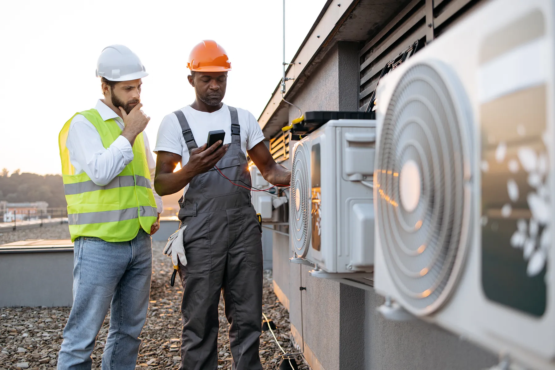hvac-technicians-testing-commercial-air-conditioning-units-sita. Two HVAC technicians inspecting outdoor air conditioning units while using a multimeter on a commercial rooftop.