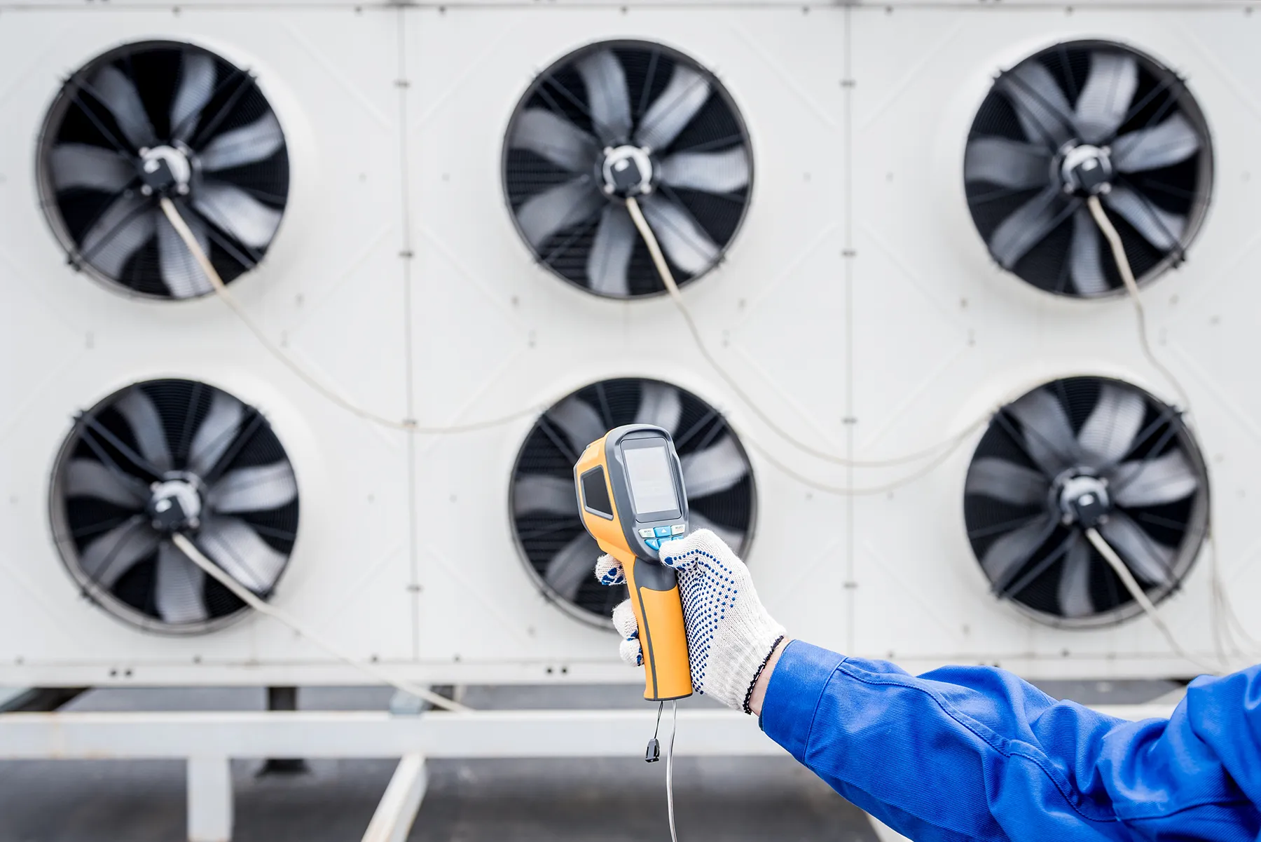 technician-testing-industrial-cooling-and-ventilation-system-sita. Technician using a handheld thermal measuring device in front of large industrial ventilation and cooling fans.