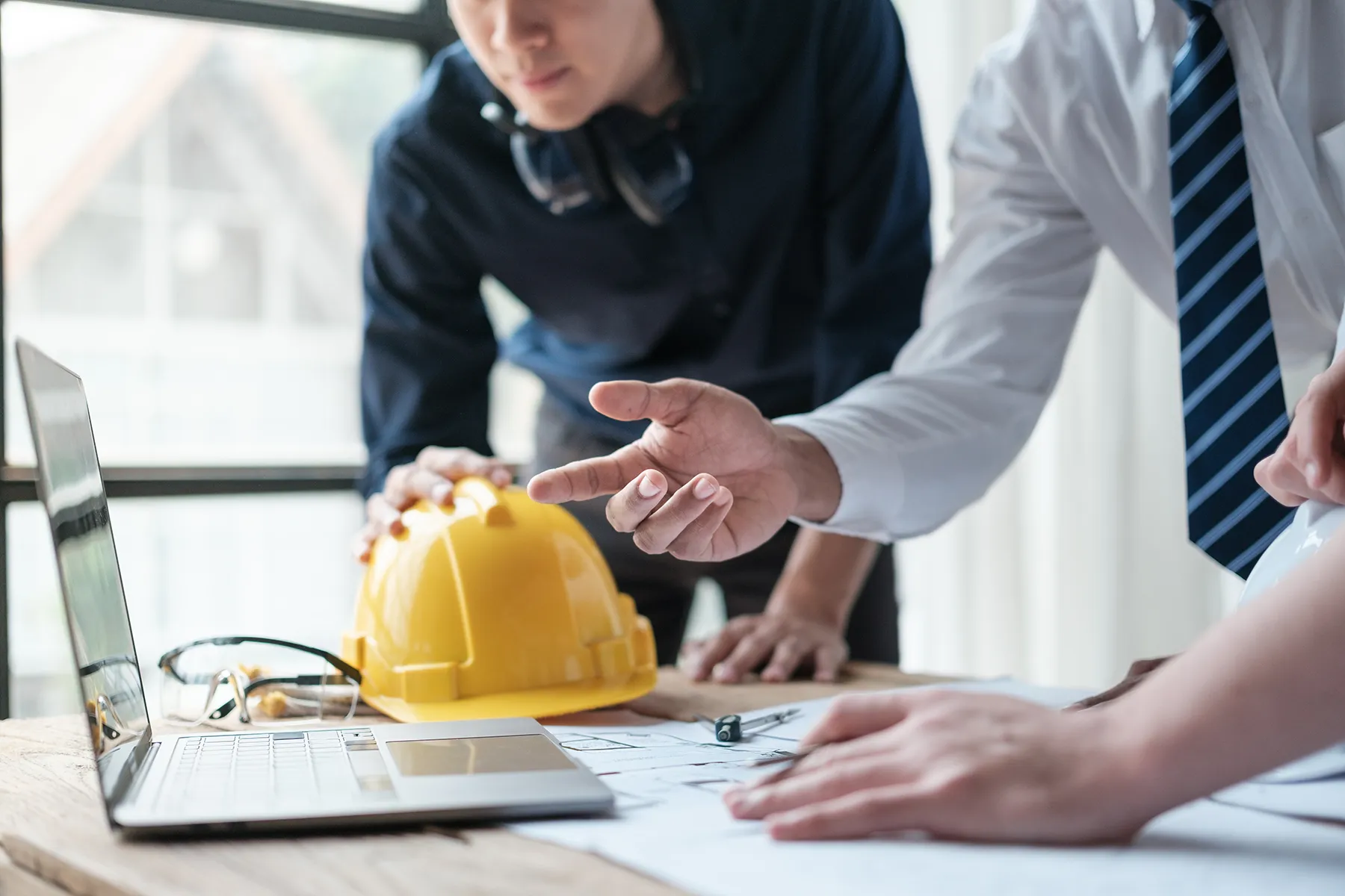 engineering-team-reviewing-construction-plans-sita. Construction and engineering professionals discussing project plans at a desk with blueprints, a laptop, and a yellow hard hat.