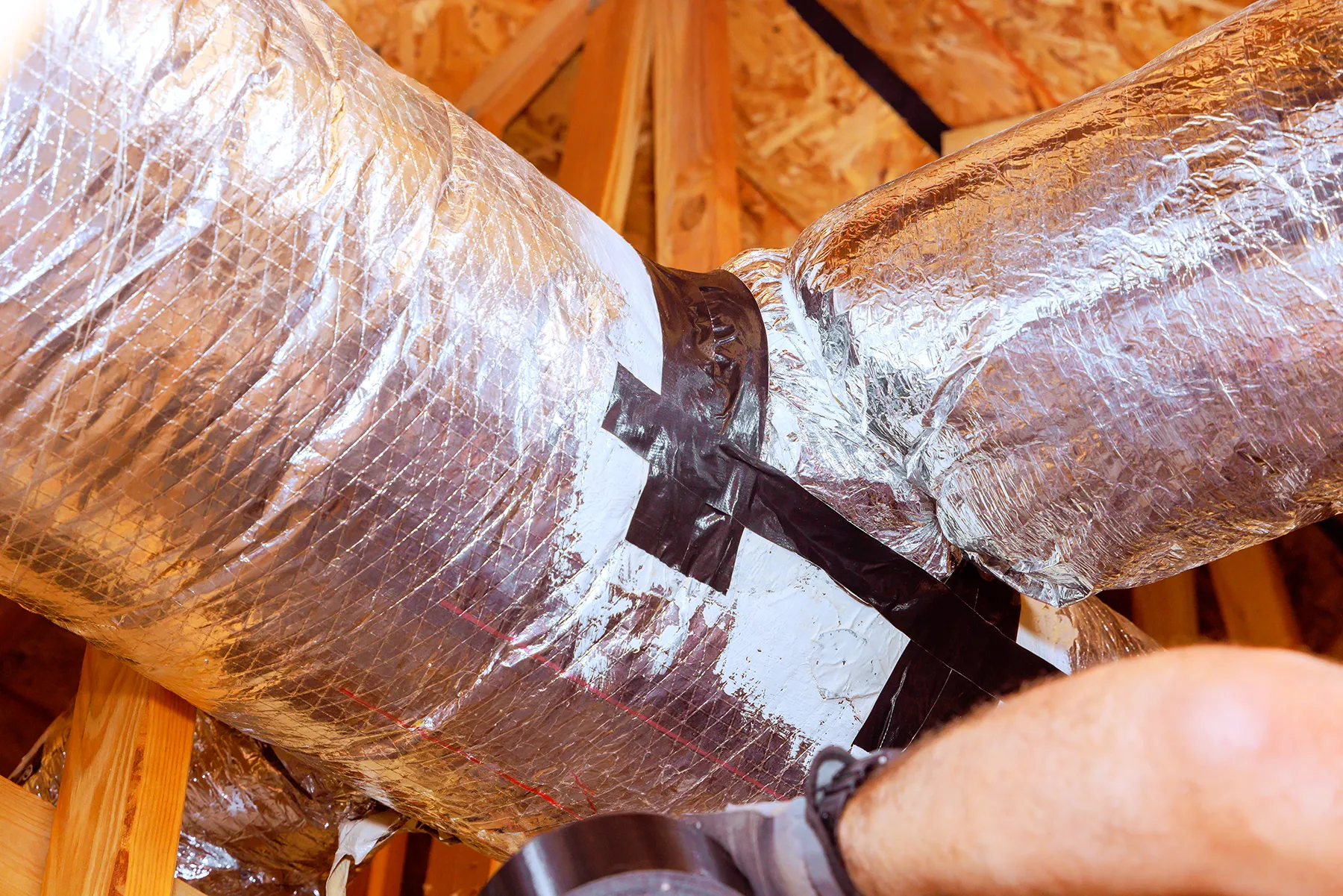 insulated-hvac-ductwork-installation-and-sealing-sita. HVAC ductwork wrapped in reflective insulation being sealed with tape during an installation or repair process in an attic space.