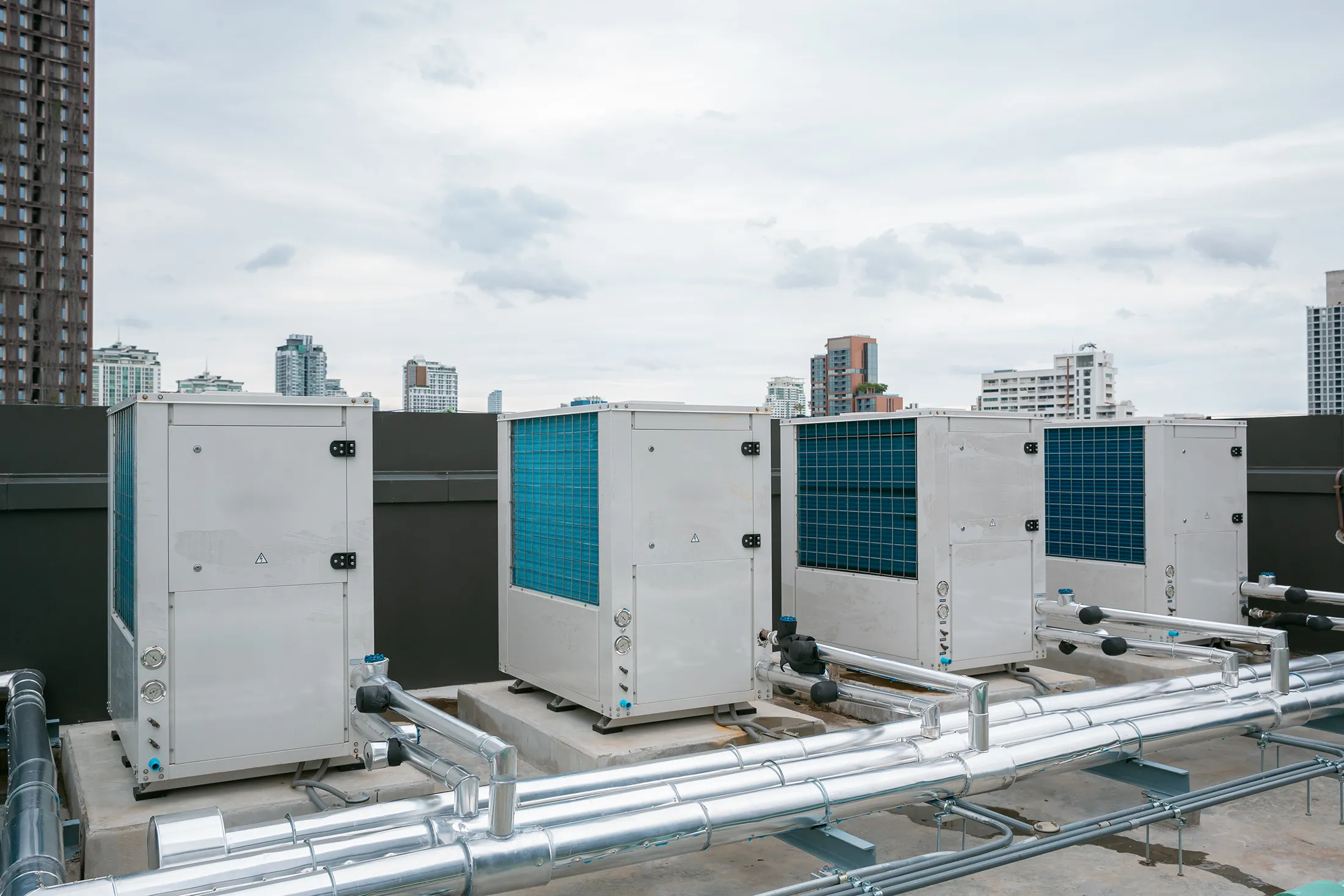 commercial-rooftop-air-conditioning-compressor-units-sita. Commercial rooftop air conditioning compressor units connected to metal piping with a city skyline in the background.