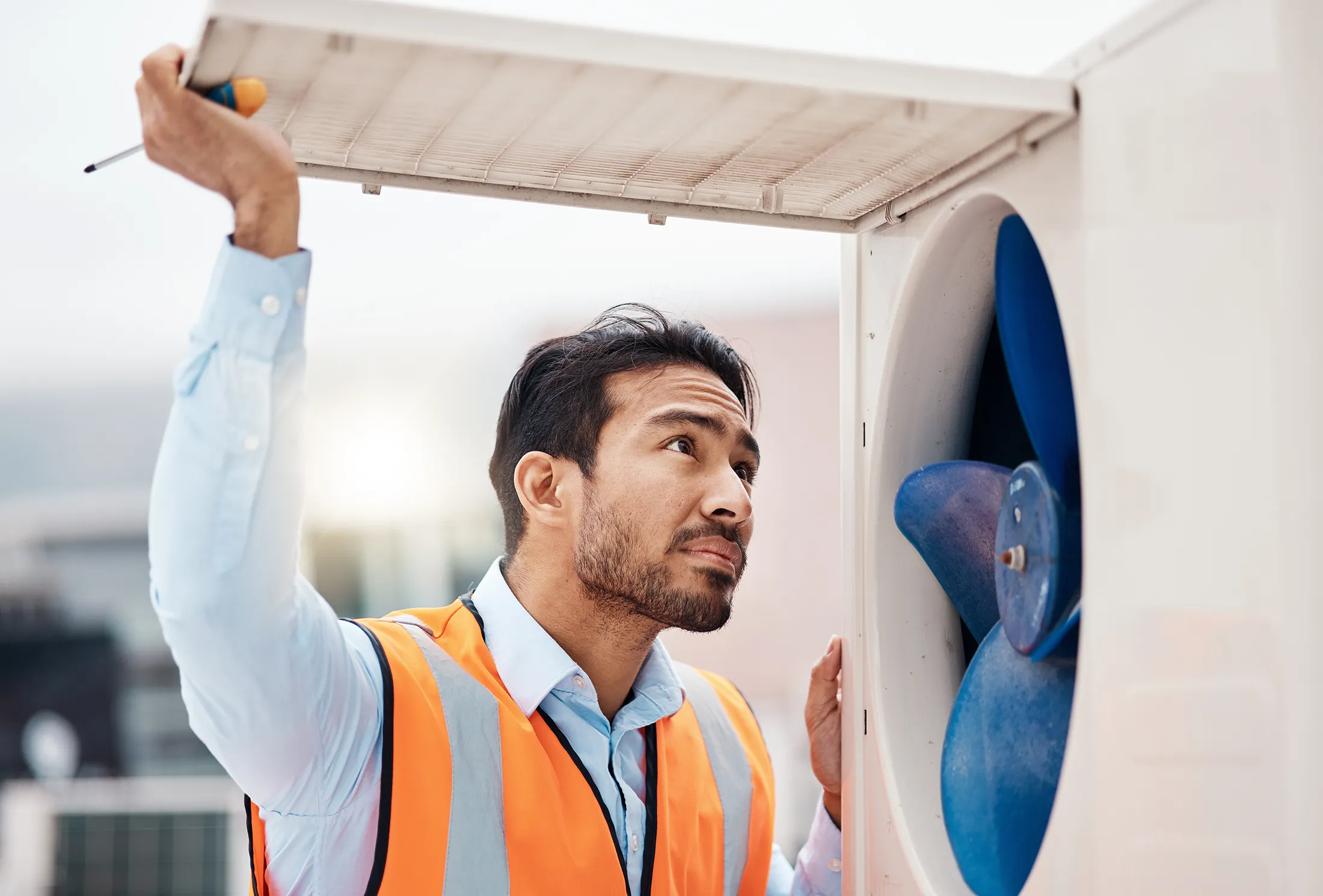 hvac-technician-performing-air-conditioning-maintenance-sita. HVAC technician inspecting and maintaining an outdoor air conditioning unit while wearing a high-visibility safety vest.